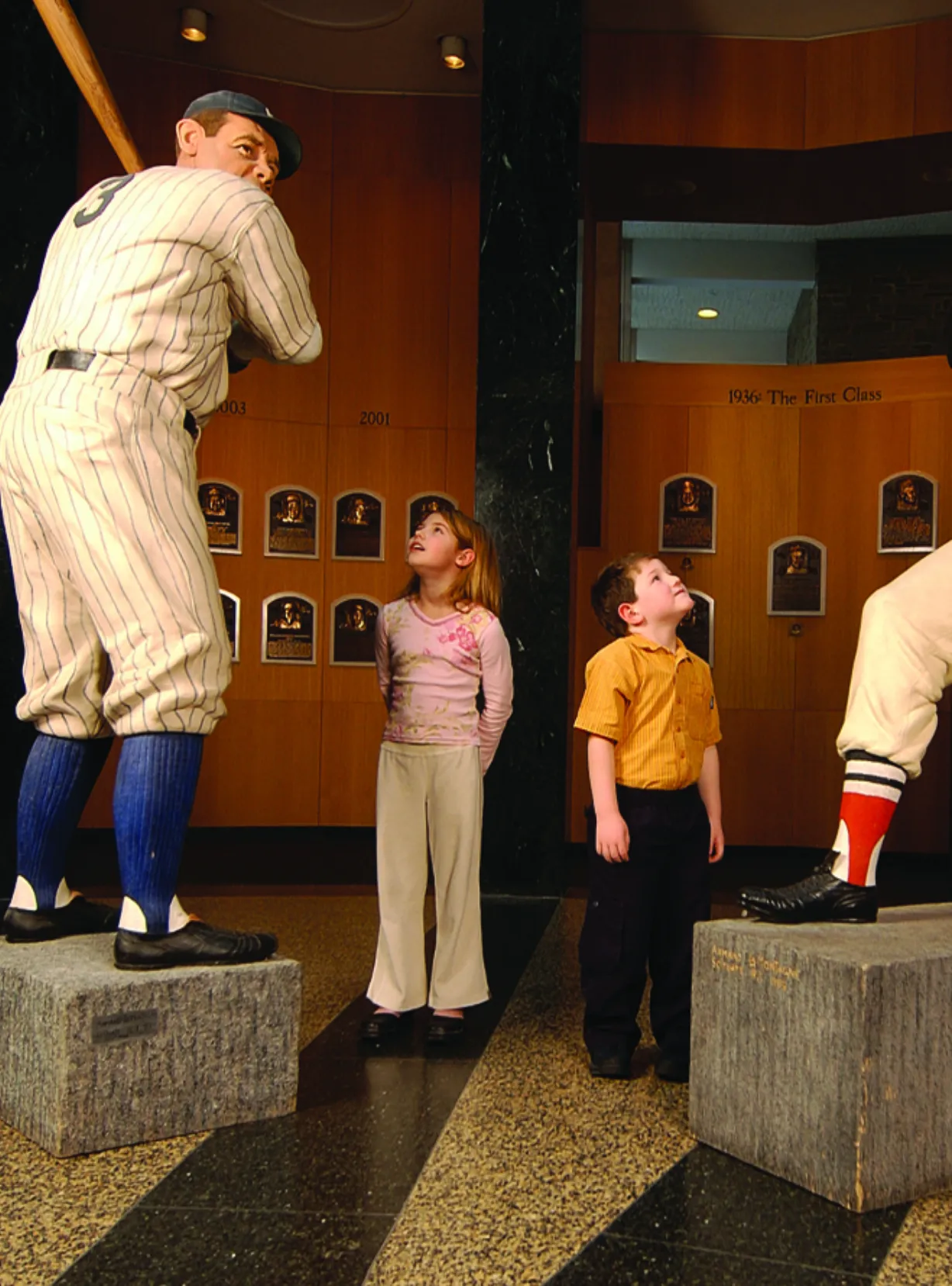 Two children admire life-sized statues of legendary baseball players at the National Baseball Hall of Fame, surrounded by plaques commemorating inductees from various years.